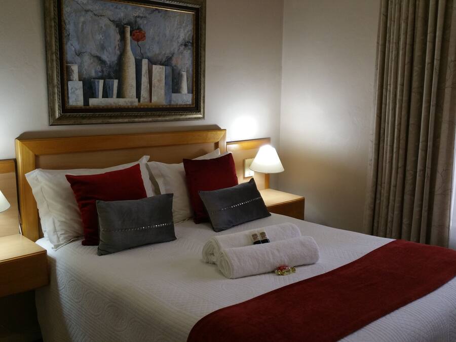 A neatly made bed with a red blanket, white pillows, and a painting above it. There are two bedside tables with lamps and a book on one.