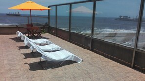 A balcony with lounge chairs and an umbrella, overlooking the ocean with ships in the distance.
