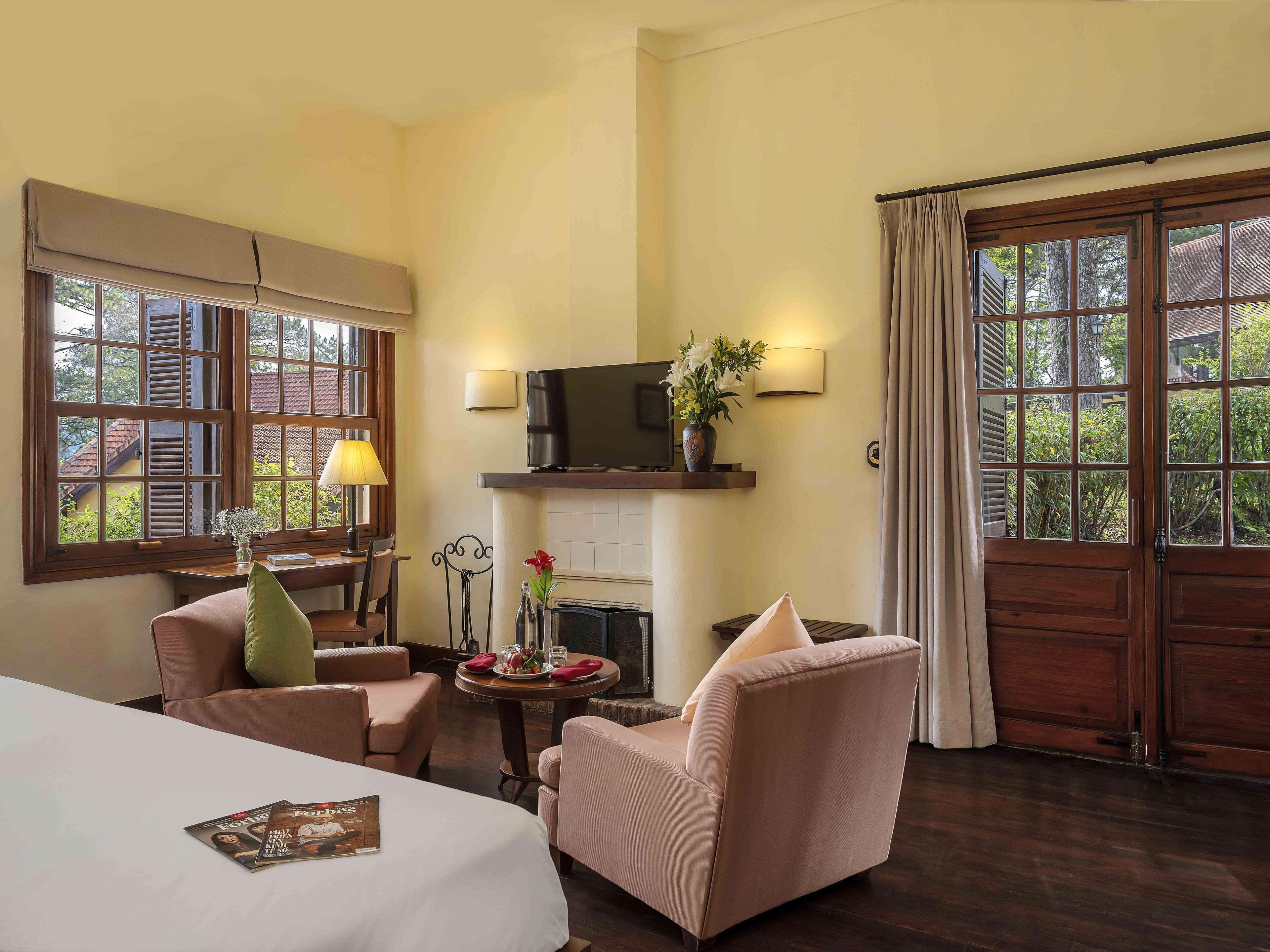 Habitación de hotel con cama, dos sillones, una mesita de noche con lámpara, televisor y chimenea.