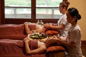 A massage therapy room with two massage tables, one occupied by a client and another with a therapist. A tray with massage oils and a bowl of fruits is on the table.