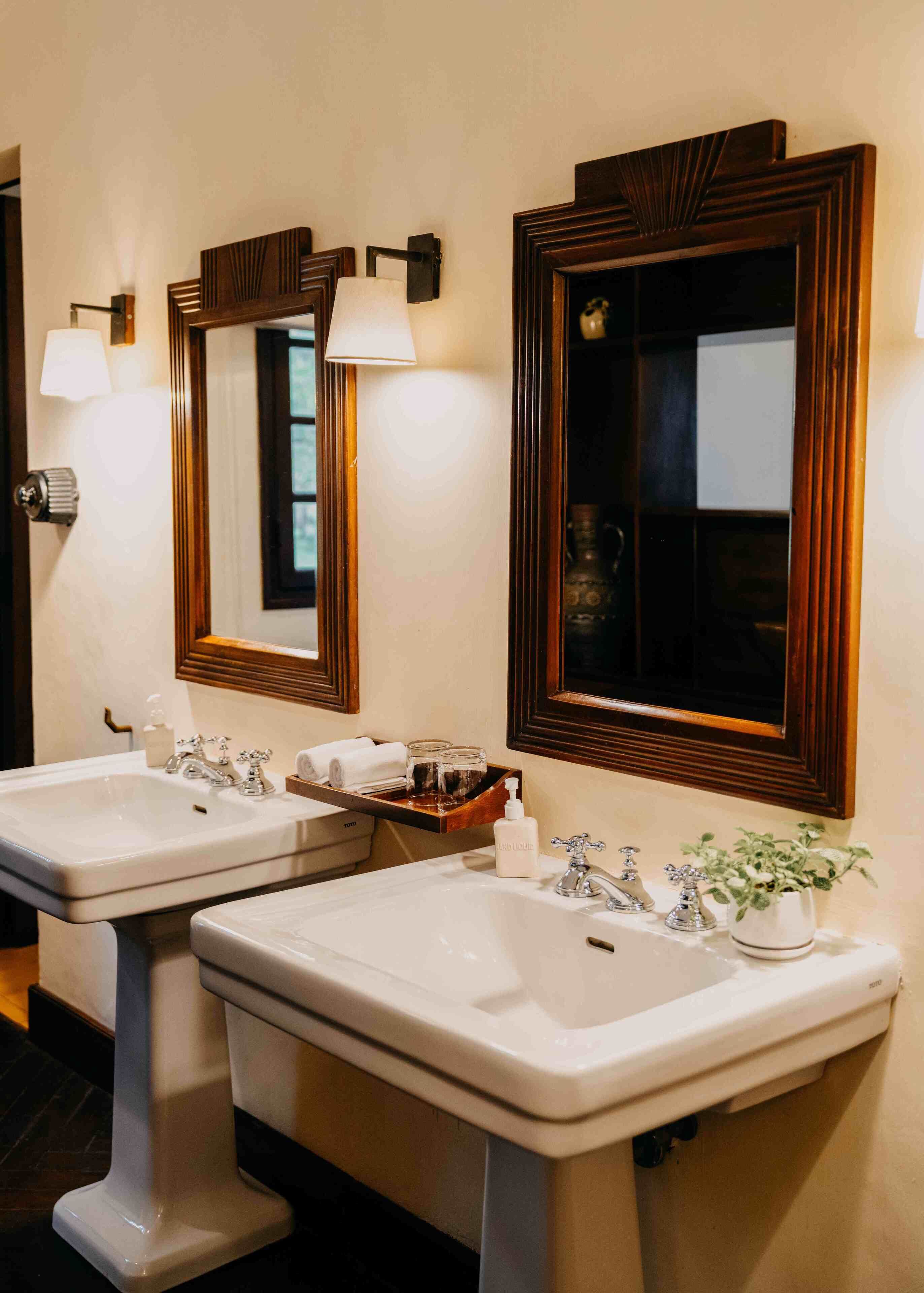 A bathroom with two sinks, each with a mirror above. There are wall-mounted lights, a towel rack, and a potted plant.