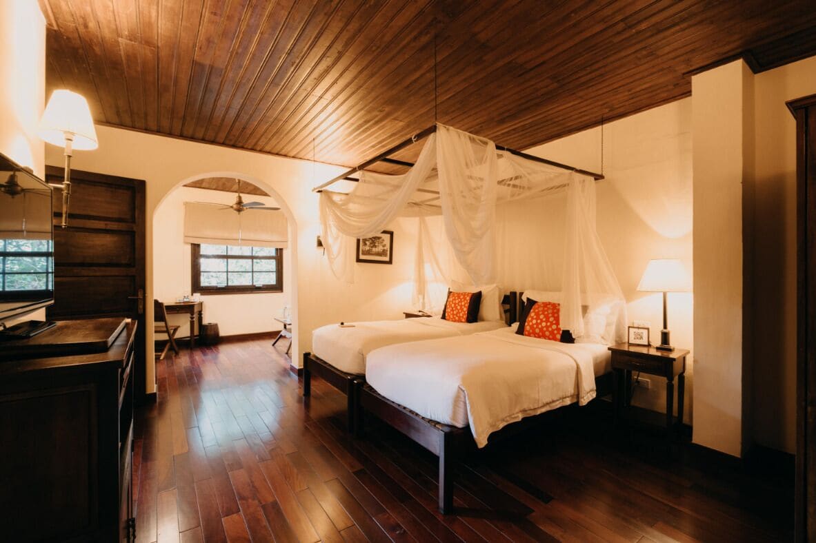 A bedroom with a canopy bed, wooden flooring, and a desk with a lamp.