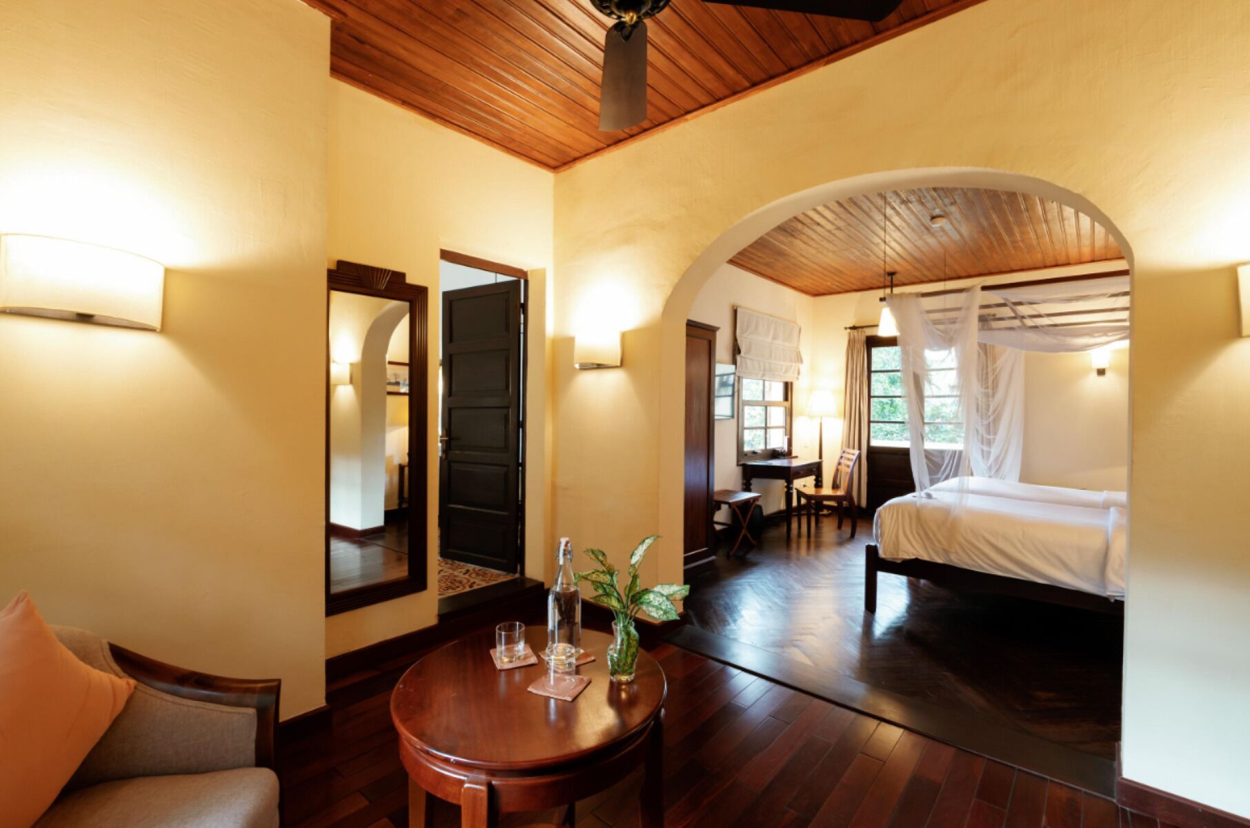 A hotel room with a wooden ceiling, a bed with a canopy, a desk with a chair, a round table with a vase and glasses, and a mirror on the wall.