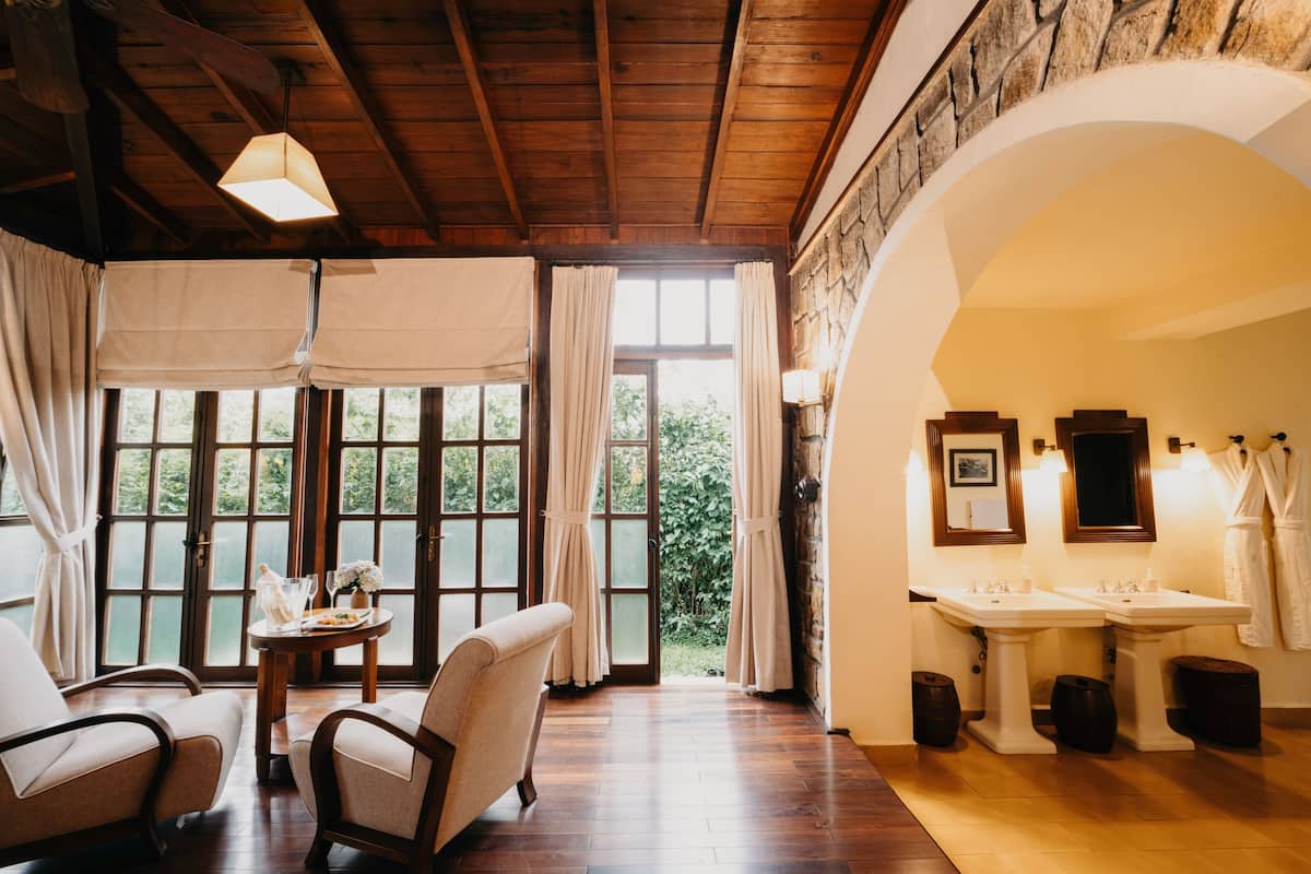 Una habitación con piso de madera, una mesa redonda con un florero, dos sillas, un ventanal con cortinas, una pared de piedra y un baño con dos lavamanos y un espejo.