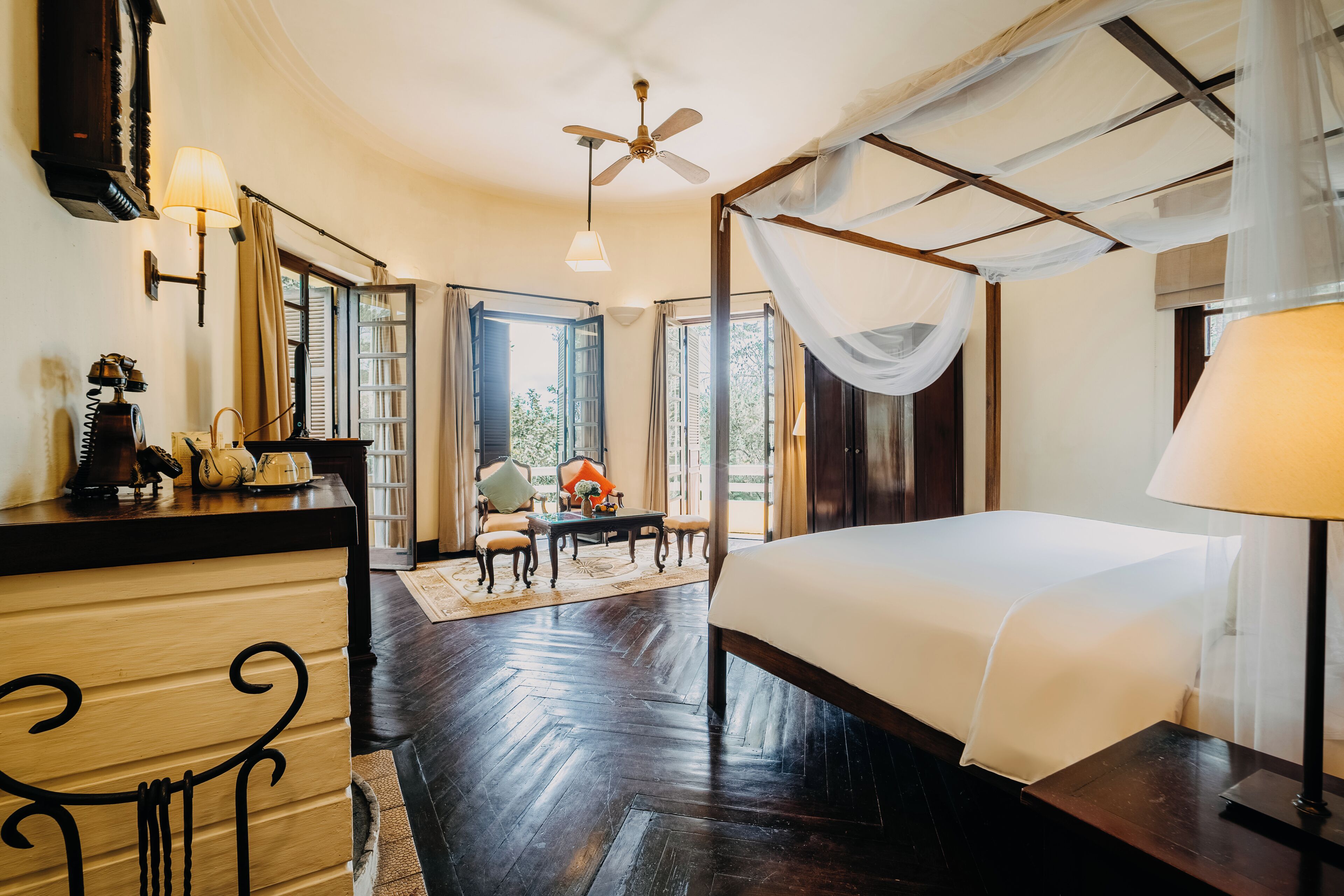 A bedroom with a four-poster bed, a wooden dresser, a desk with chairs, and a view of the outdoors through large windows.