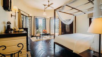 A bedroom with a four-poster bed, a wooden dresser, a desk with chairs, and a view of the outdoors through large windows.