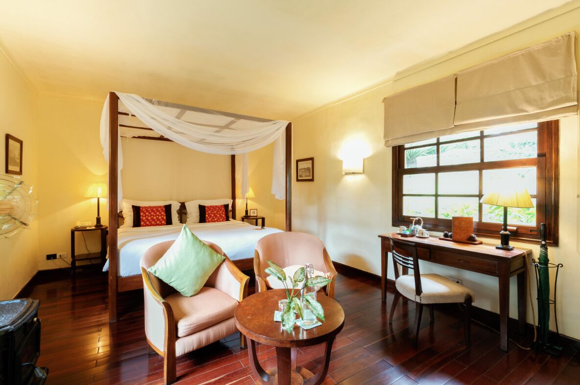 A bedroom with a four-poster bed, a desk with a chair, a small round table with a vase, and a window with a view of greenery.