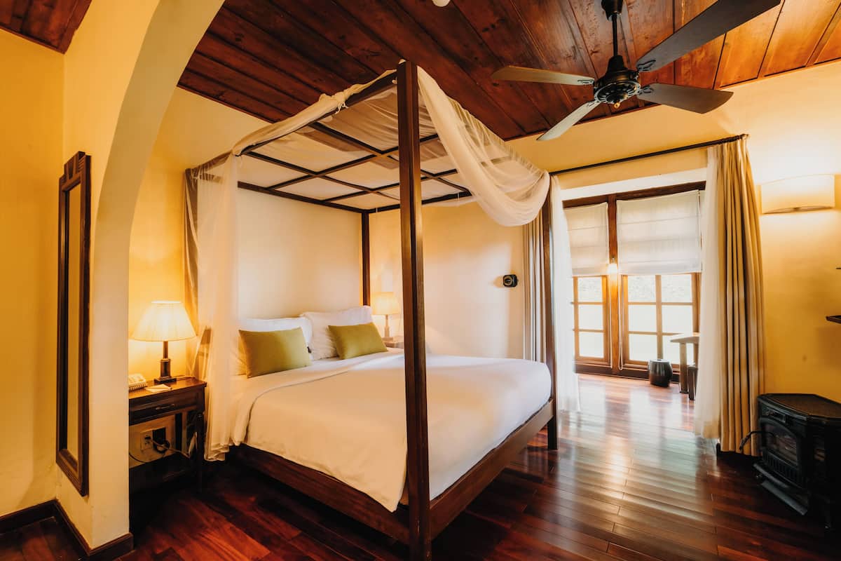 Una cama con dosel y columnas, suelo de madera, ventilador de techo y vistas al exterior a través de un gran ventanal.