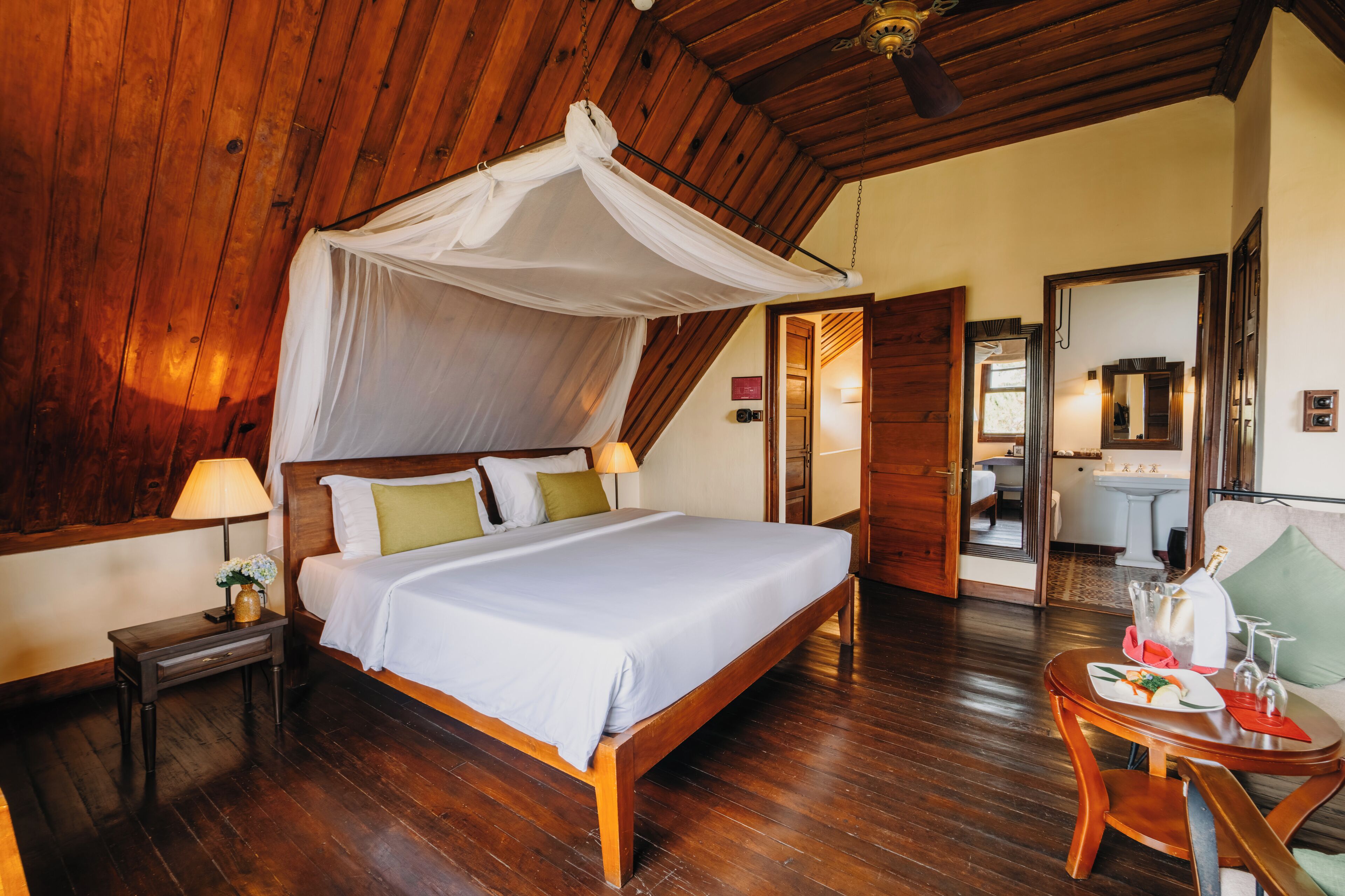 A wooden-floored bedroom with a canopy bed, bedside table with a lamp, and a small table with a bottle and glasses.