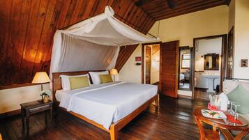 A wooden-floored bedroom with a canopy bed, bedside table with a lamp, and a small table with a bottle and glasses.