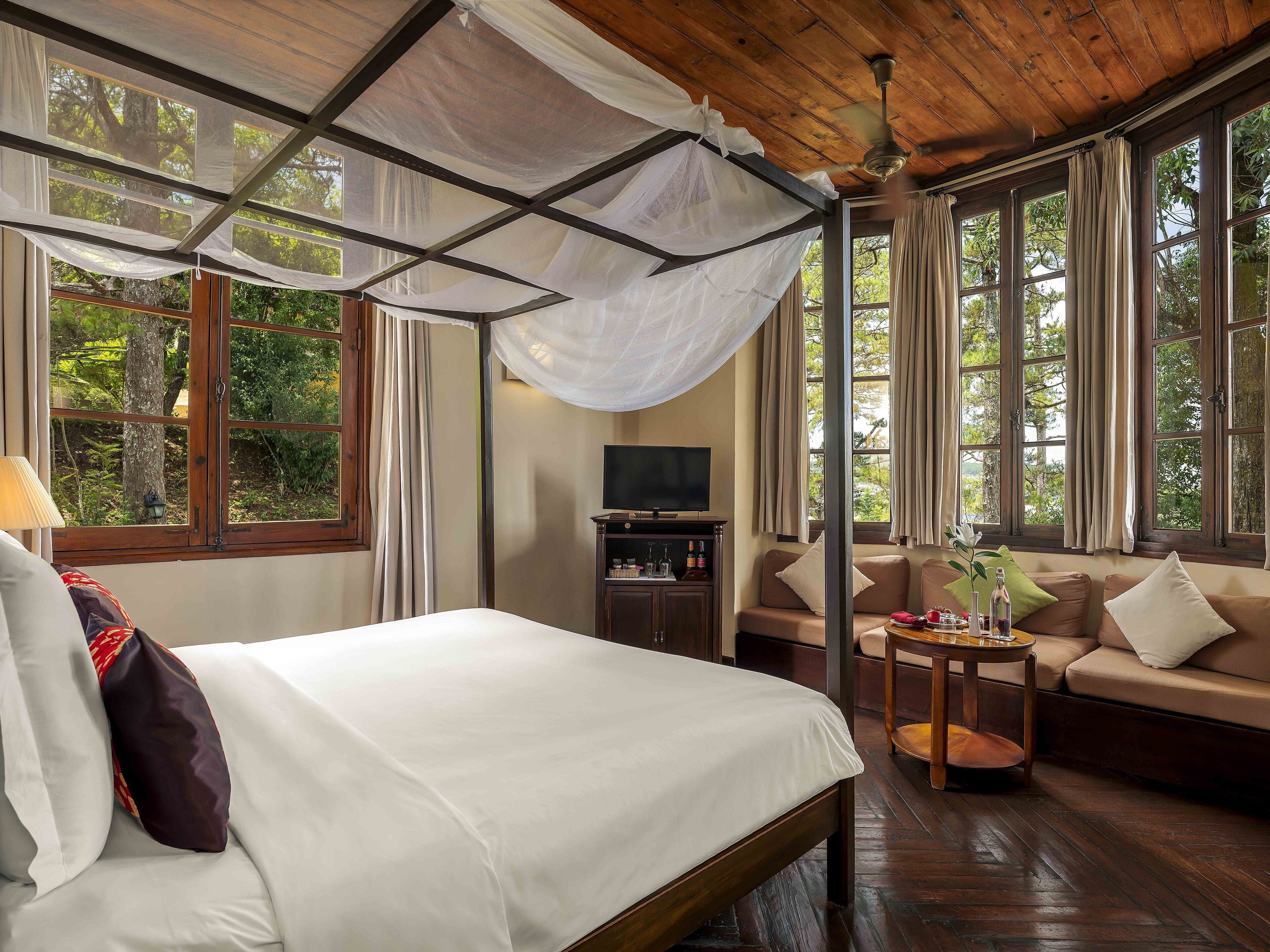 A bedroom with a bed, a TV, a sofa, and large windows.