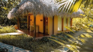 A thatched-roof bungalow with a wooden porch, surrounded by lush greenery and palm trees.