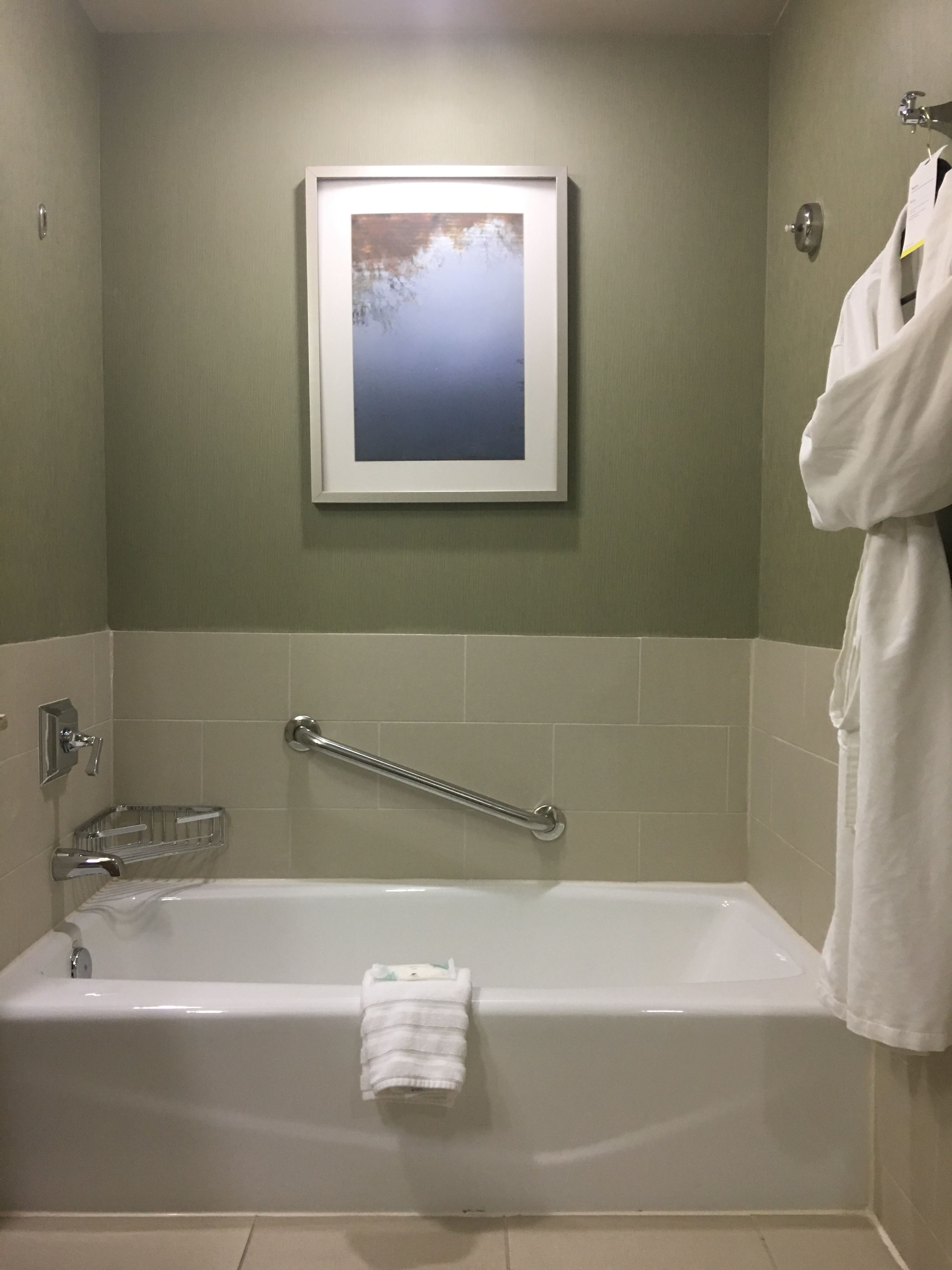 Ein Badezimmer mit einer Badewanne, einer Handstange, einem gerahmten Bild an der Wand und einem Wandtuch.