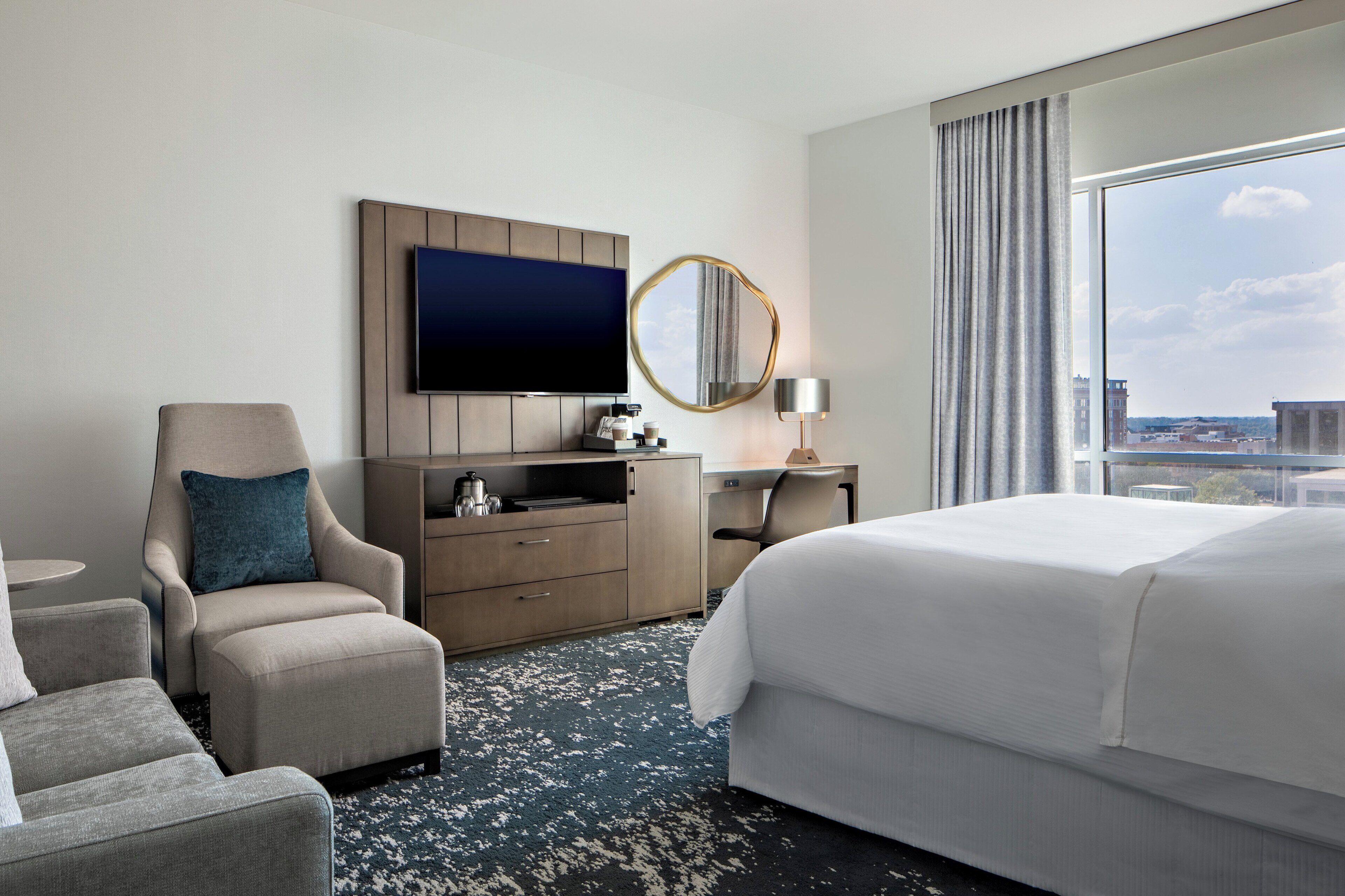 A hotel room with a large bed, a desk with a flat-screen TV, a chair, a dresser, and a view of the city through a large window.