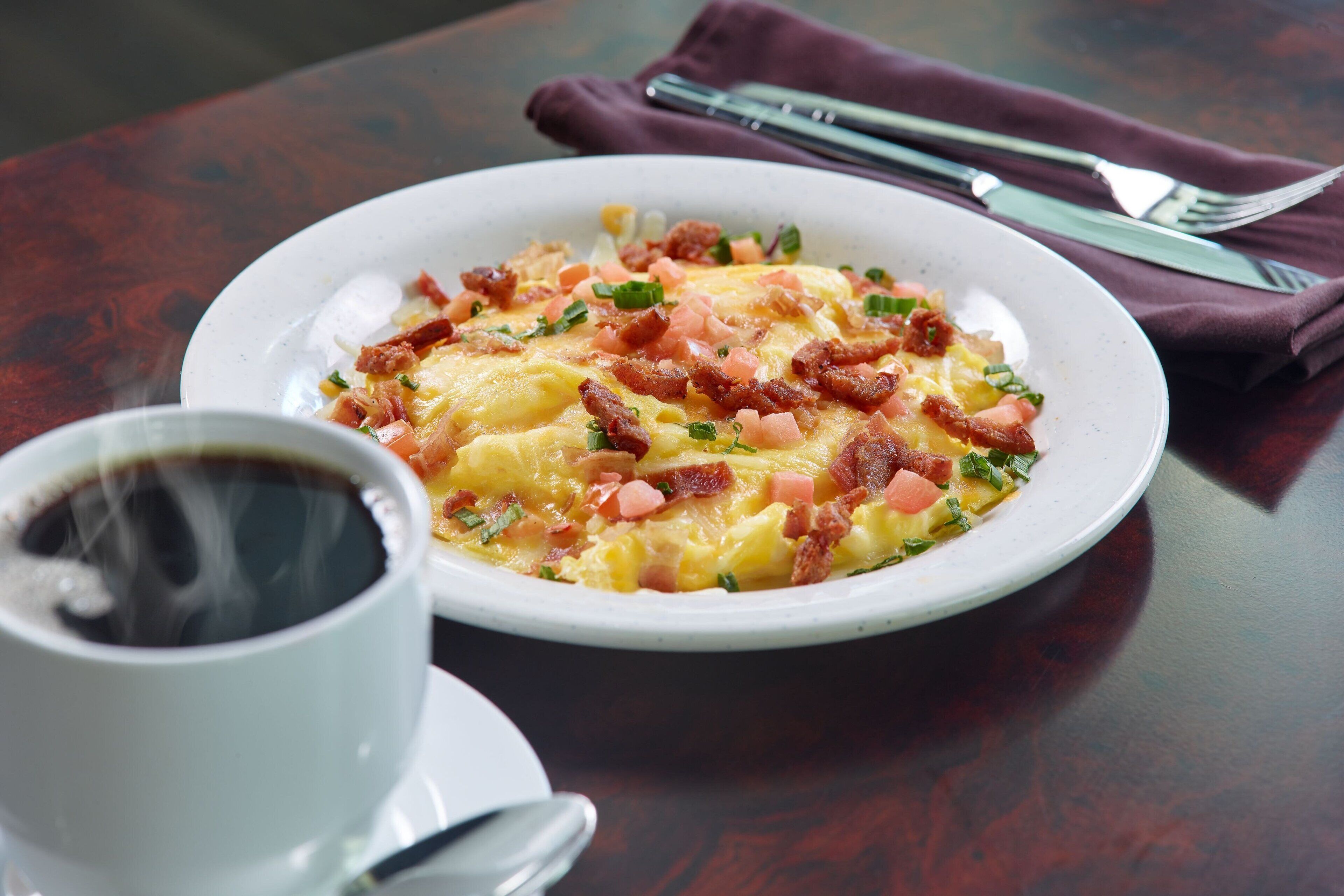 Une assiette d’œufs brouillés, de lardons et de tomates coupées en dés, accompagnée d’une tasse de café sur une table.