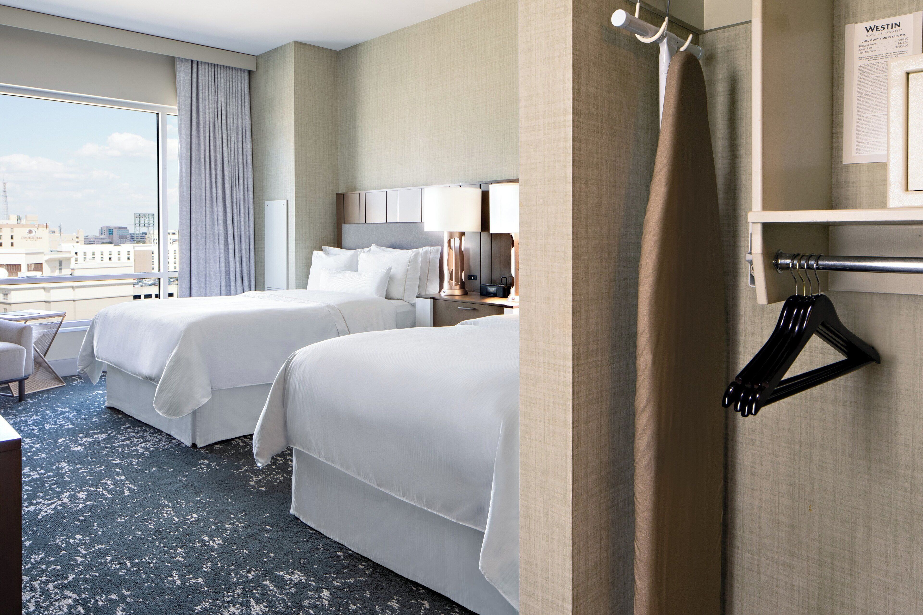 A hotel room with two beds, a desk, a chair, a TV, and a view of the city.