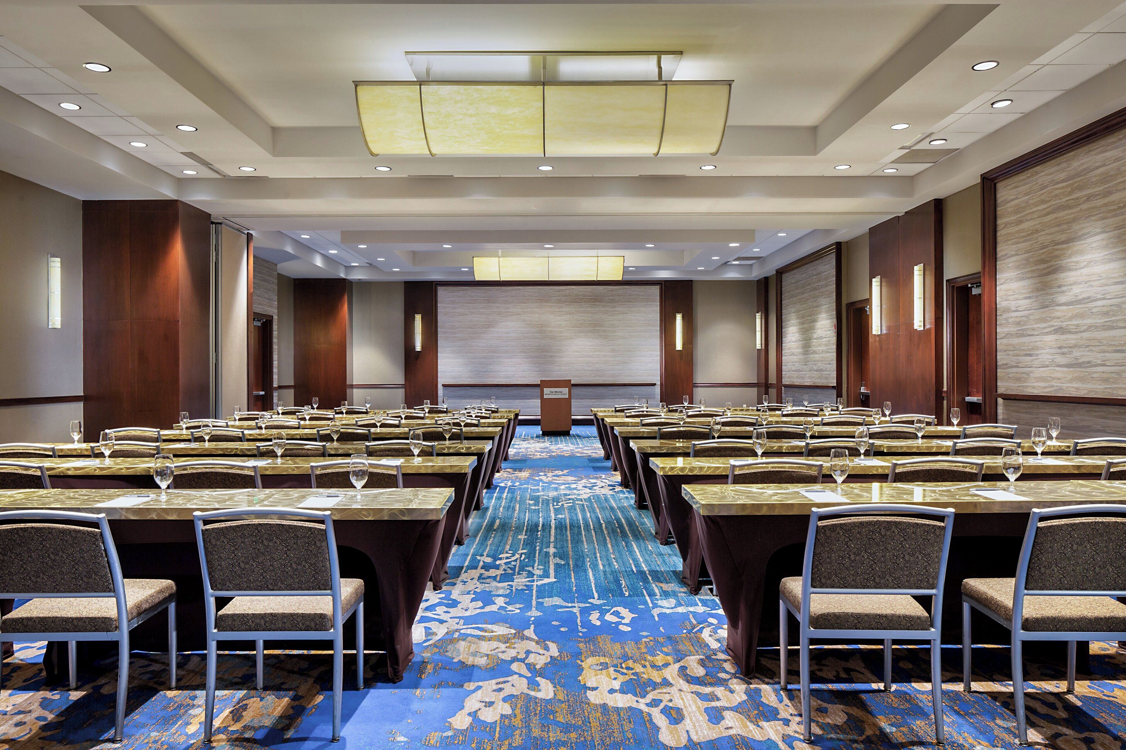 A conference room with rows of tables and chairs, a podium, and a large screen at the front.