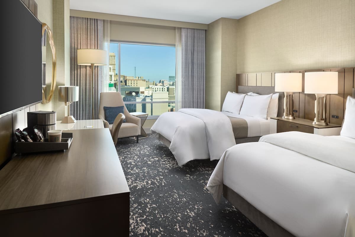 A hotel room with two beds, a desk, a chair, a TV, and a large window offering a city view.