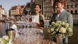 Una pareja de pie frente a una mesa con copas y botellas de vino, con un paisaje urbano de fondo.