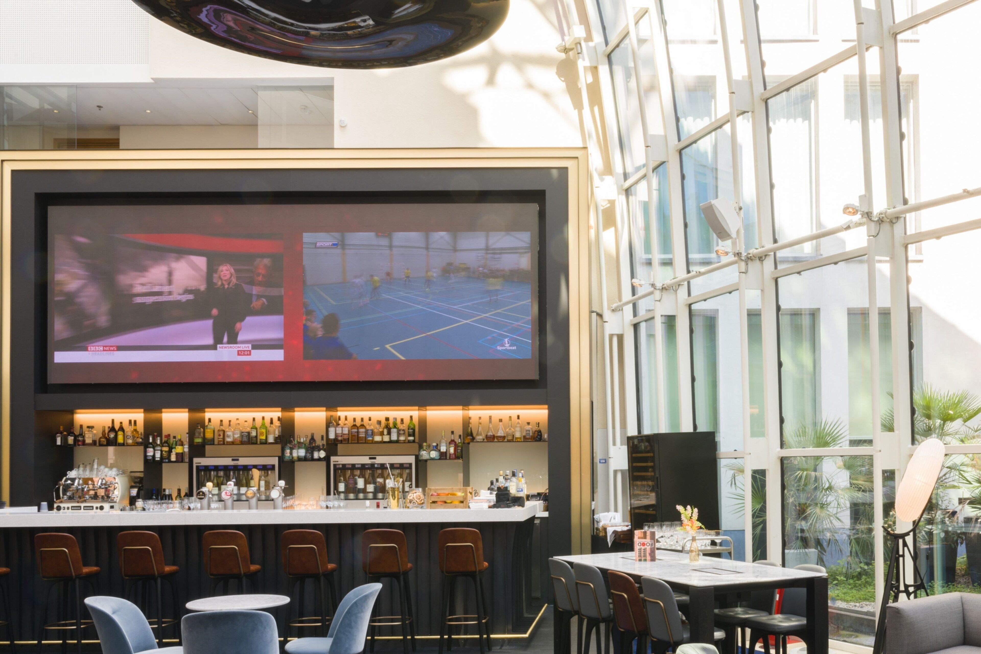 A modern bar area with a large TV screen, bar stools, and a variety of bottles.