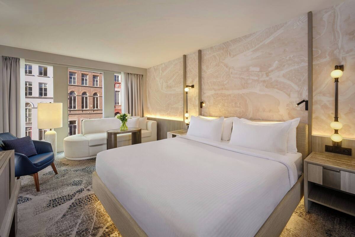 Habitación de hotel con una cama grande, un escritorio, una silla y vistas a edificios a través de las ventanas.