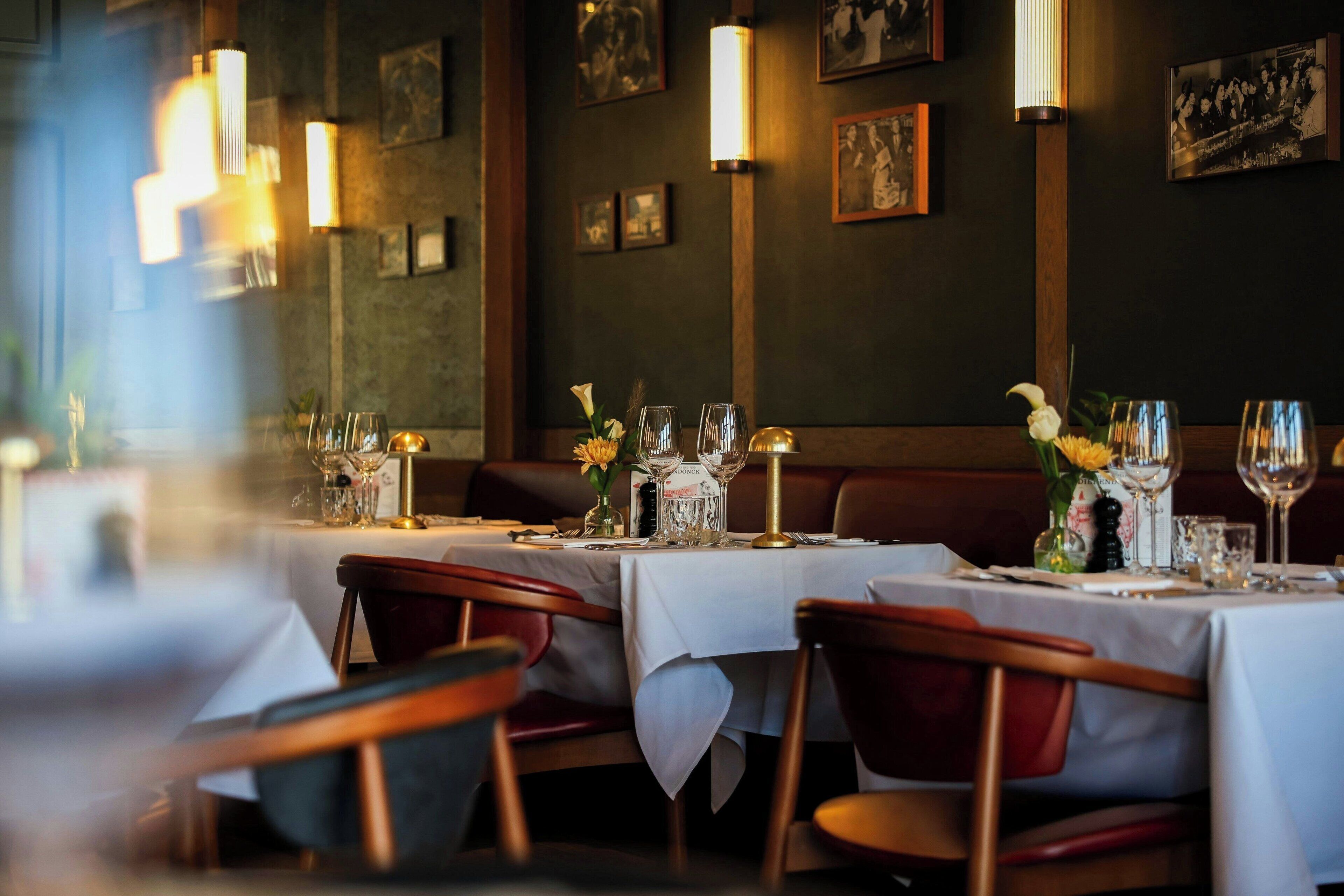 A restaurant interior with tables set for dining, featuring a dark wall with framed pictures and wall-mounted lights.