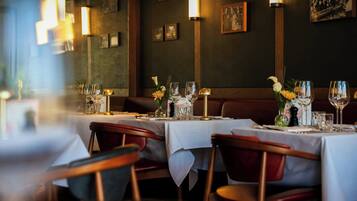 A restaurant interior with tables set for dining, featuring a dark wall with framed pictures and wall-mounted lights.