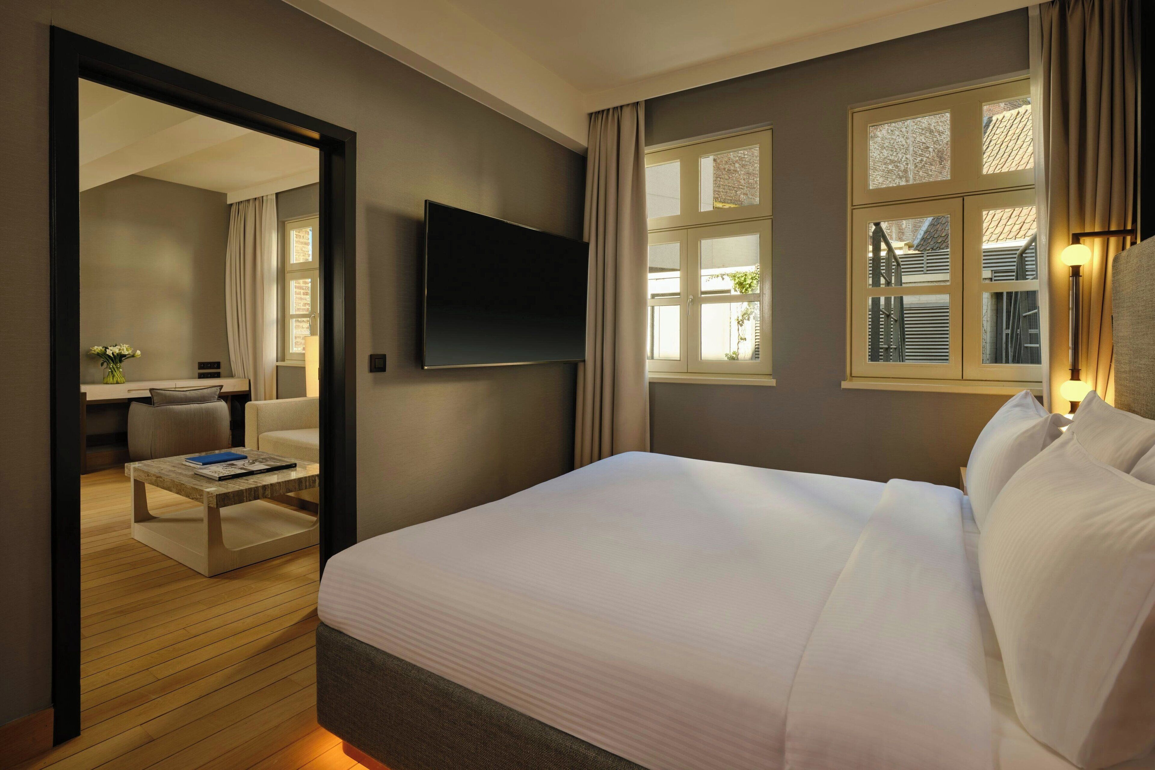Habitación de hotel con una cama grande, un televisor de pantalla plana montado en la pared y vistas al exterior a través de una ventana con cortinas.
