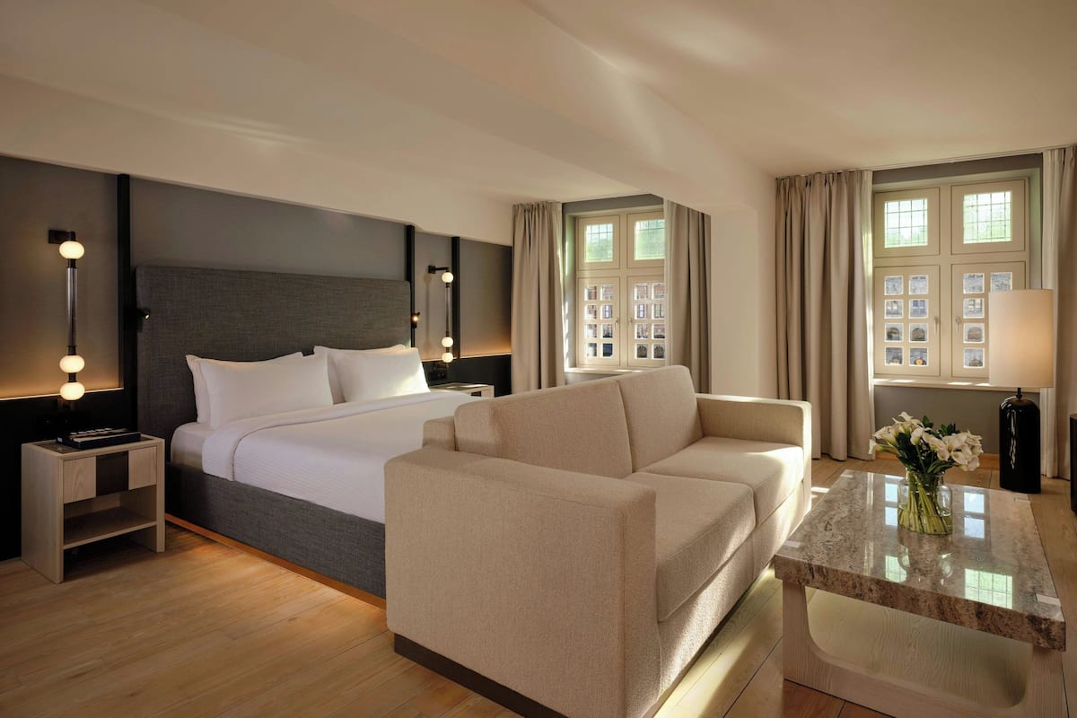 Una habitación de hotel con cama, sofá, mesita de noche con lámpara, mesa con un jarrón de flores y ventana con cortinas.