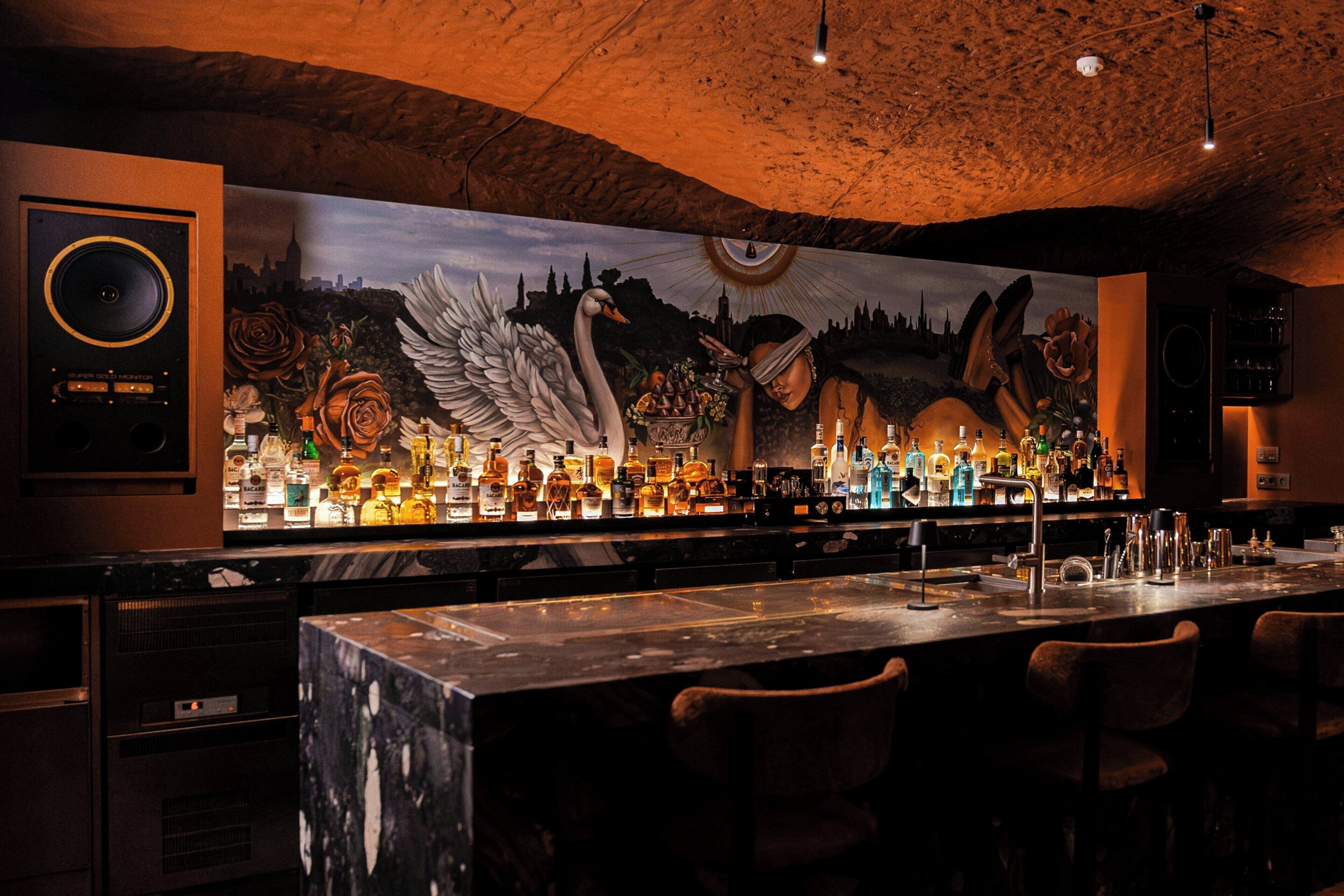 A bar with a large mural, bar stools, and a variety of bottles.