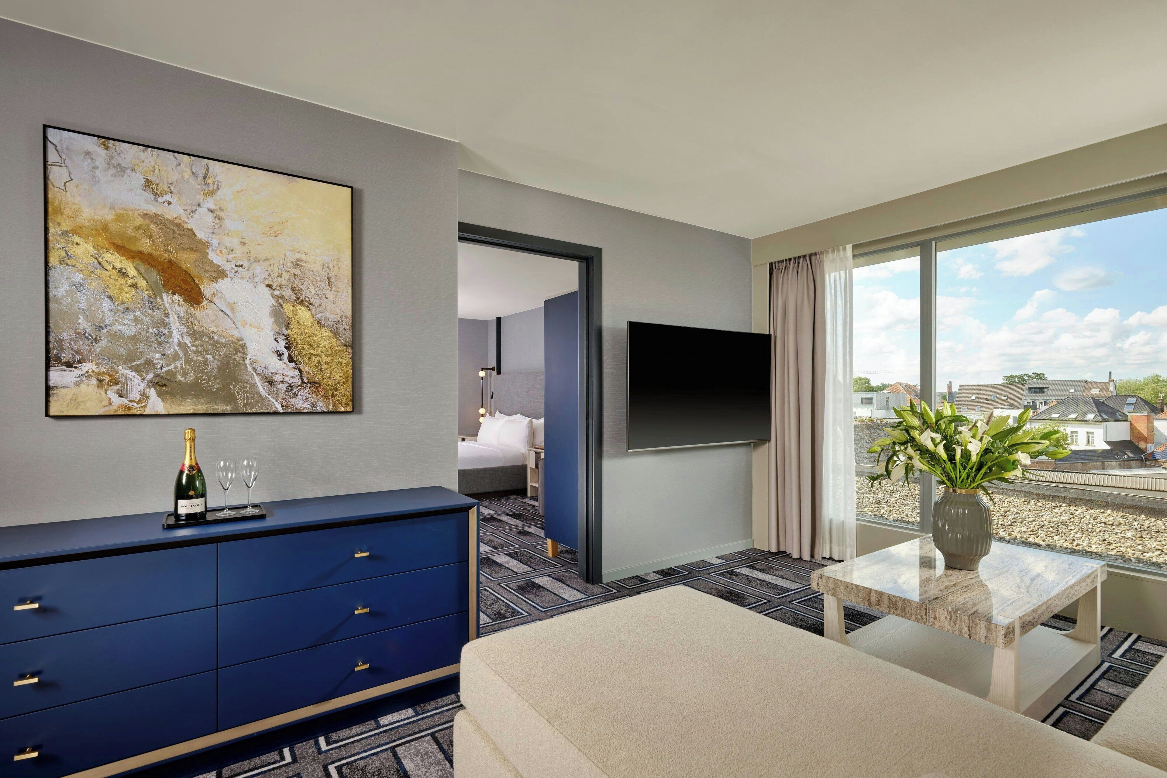 Habitación de hotel moderna con un cuadro abstracto grande, un minibar, un televisor de pantalla plana, un ventanal con vistas a la ciudad y una pequeña mesa con un florero.