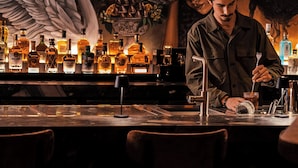 Un bar con un cantinero preparando una bebida, botellas de licores y un mural con un cisne y un paisaje urbano.
