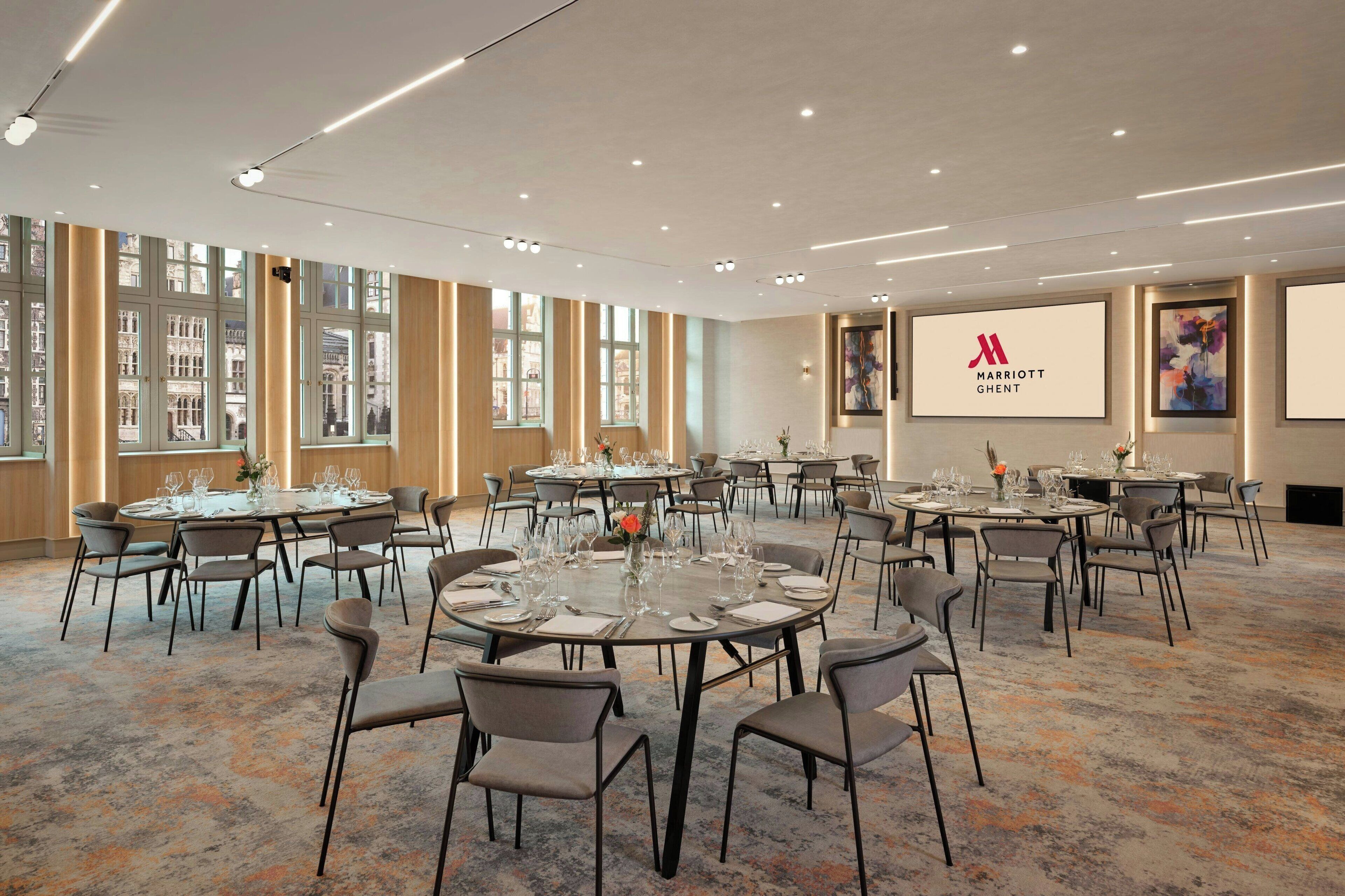 Een conferentiezaal van een Marriott hotel met meerdere ronde tafels, stoelen en een groot scherm waarop het Marriott logo staat.