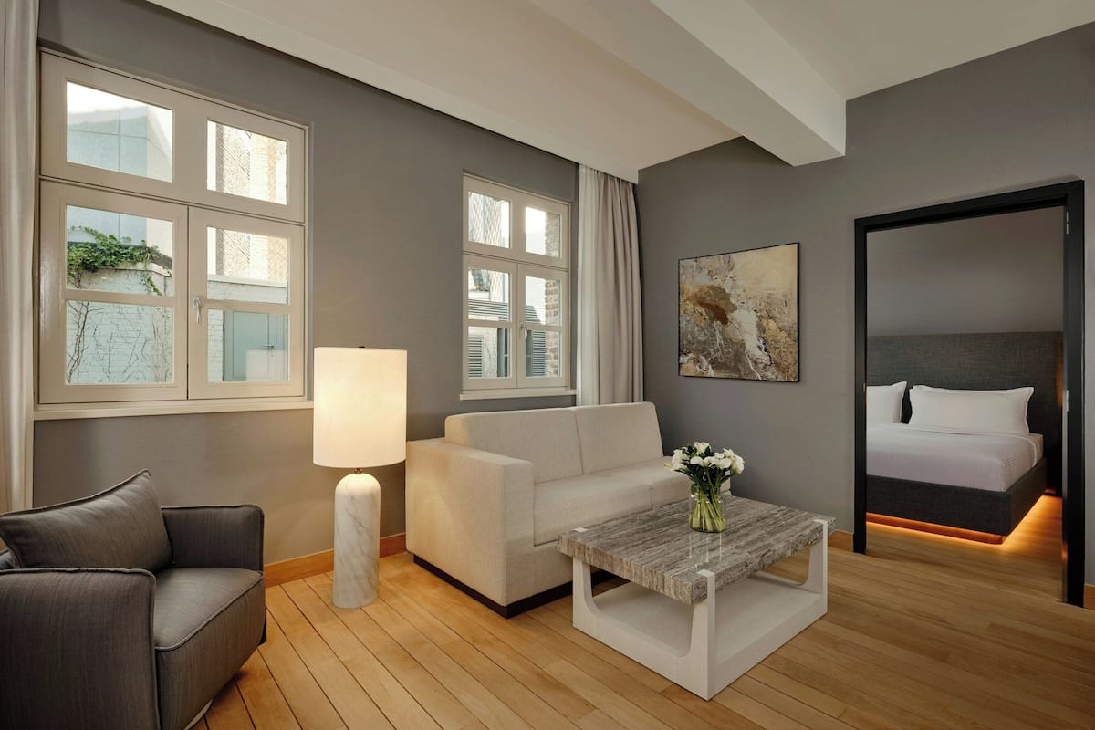 Una habitación de hotel moderna con un sofá gris, un sillón blanco, una mesa de centro de madera y una cama con cabecero.