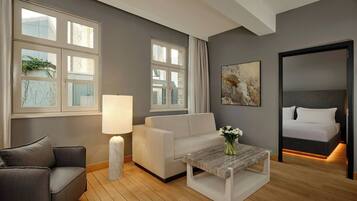 A modern hotel room with a grey sofa, a white armchair, a wooden coffee table, and a bed with a headboard.