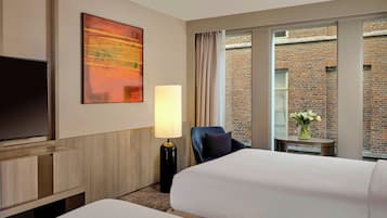 Ein Hotelzimmer mit zwei Betten, einem Fernseher, einer Lampe, einem Sessel und Blick auf ein Backsteingebäude.