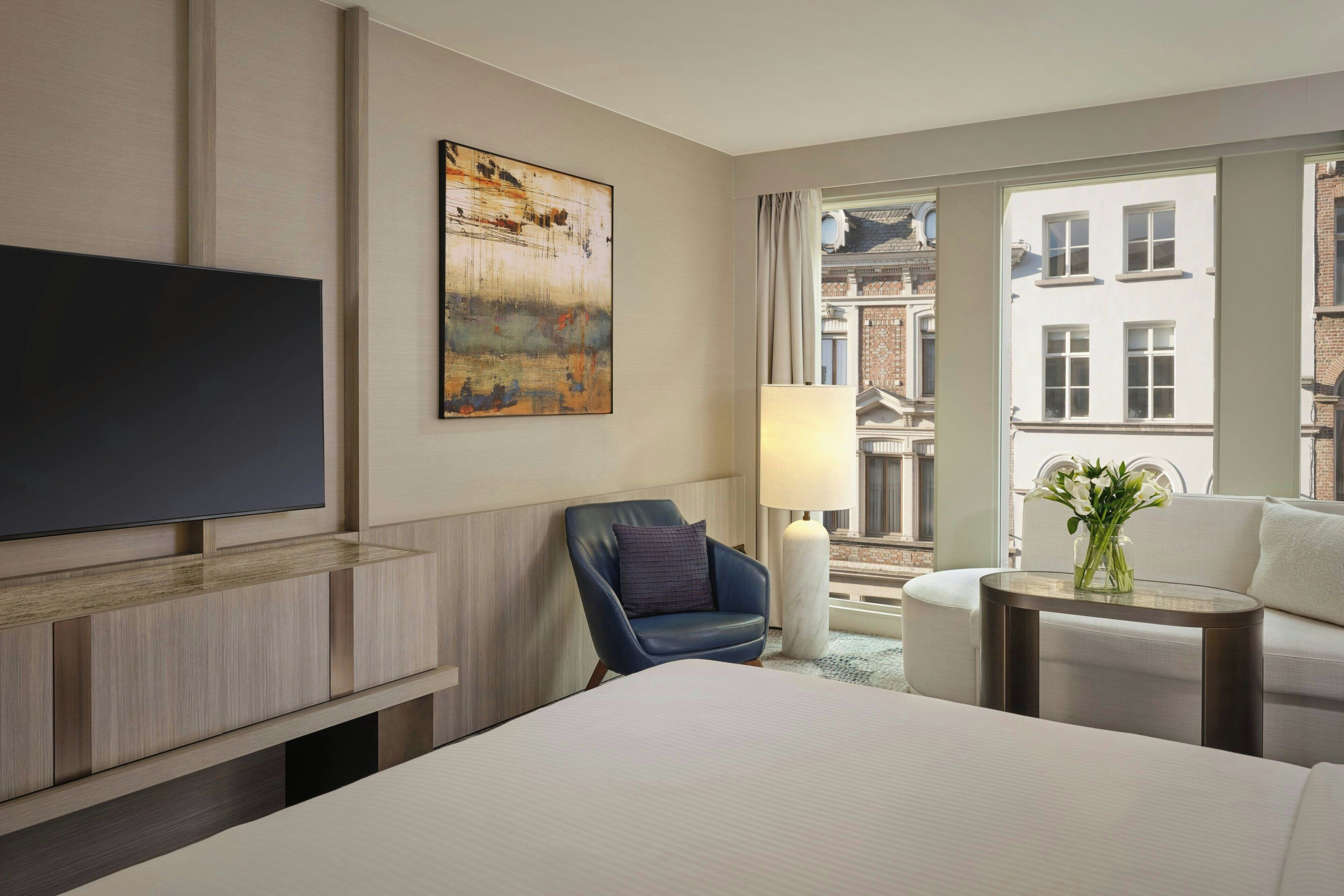 A hotel room with a bed, a flat-screen TV, a blue armchair, a small table with a lamp, and a view of buildings through a window.