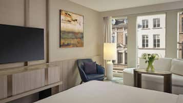 A hotel room with a bed, a flat-screen TV, a blue armchair, a small table with a lamp, and a view of buildings through a window.