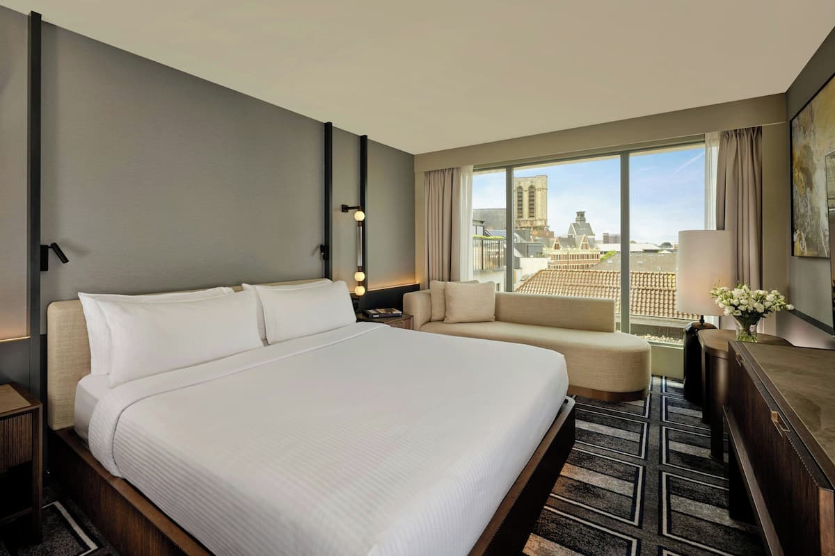 Habitación de hotel con una cama grande, un sofá, un escritorio y vistas a la ciudad a través de amplios ventanales.