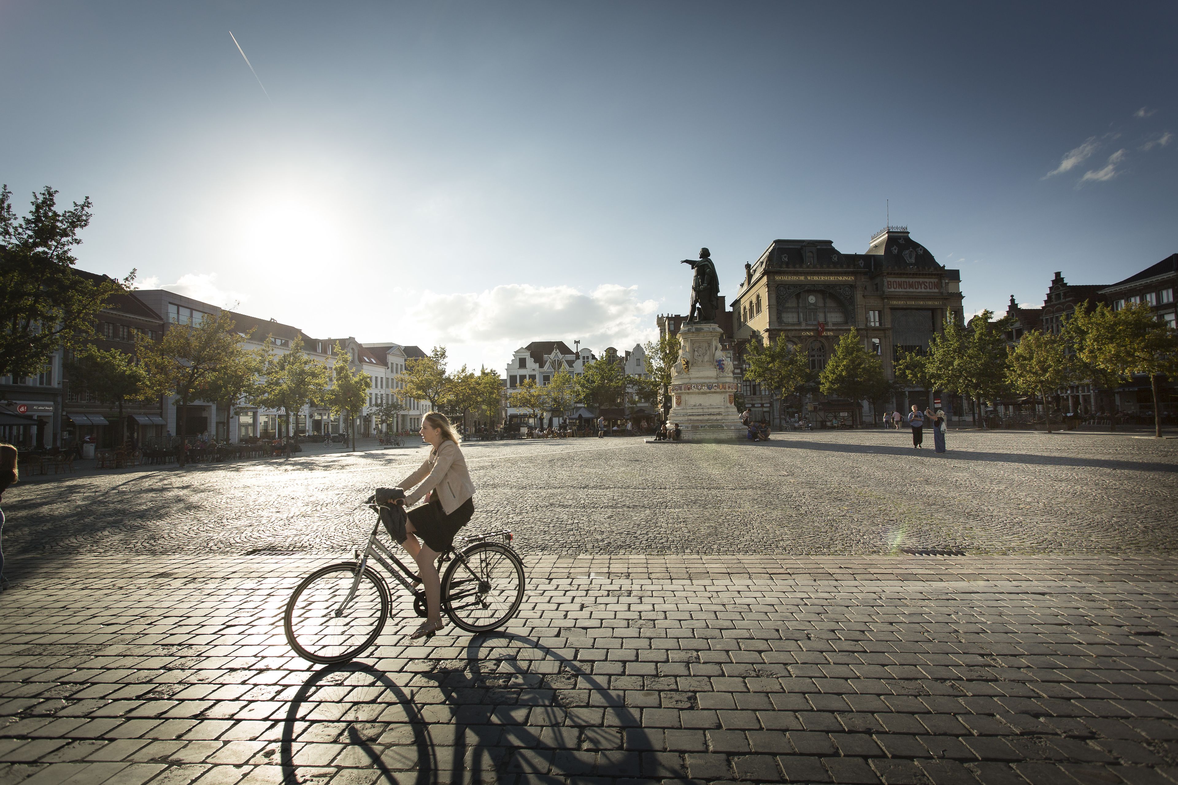 A cobblestone square with a cyclist, historic buildings, and a statue in the background.