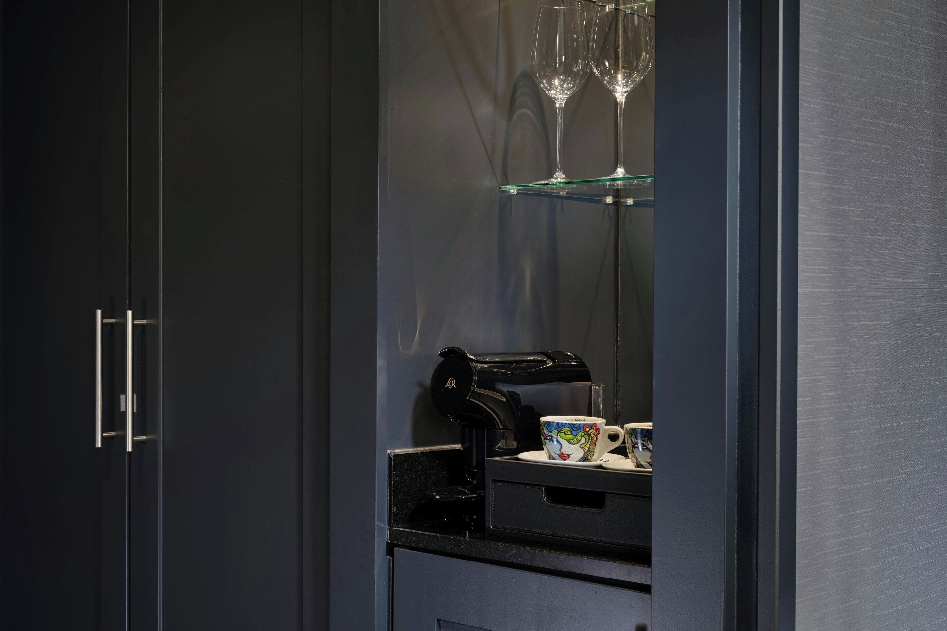 Una cocina moderna con una cafetera, una taza y una copa de vino sobre un estante de vidrio.