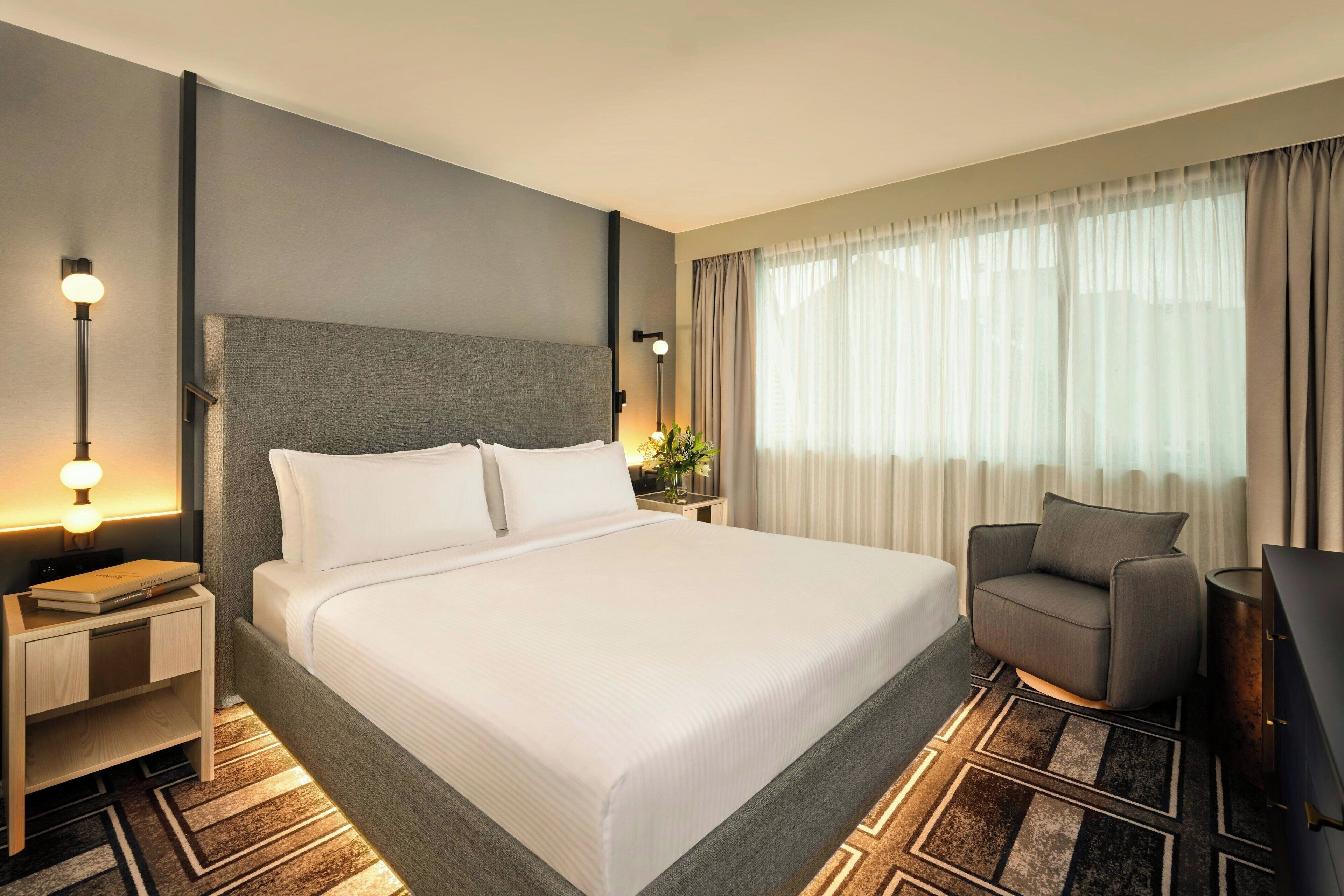 Une chambre d’hôtel moderne avec un grand lit, une table de chevet avec une lampe, une chaise et une grande fenêtre avec des rideaux.
