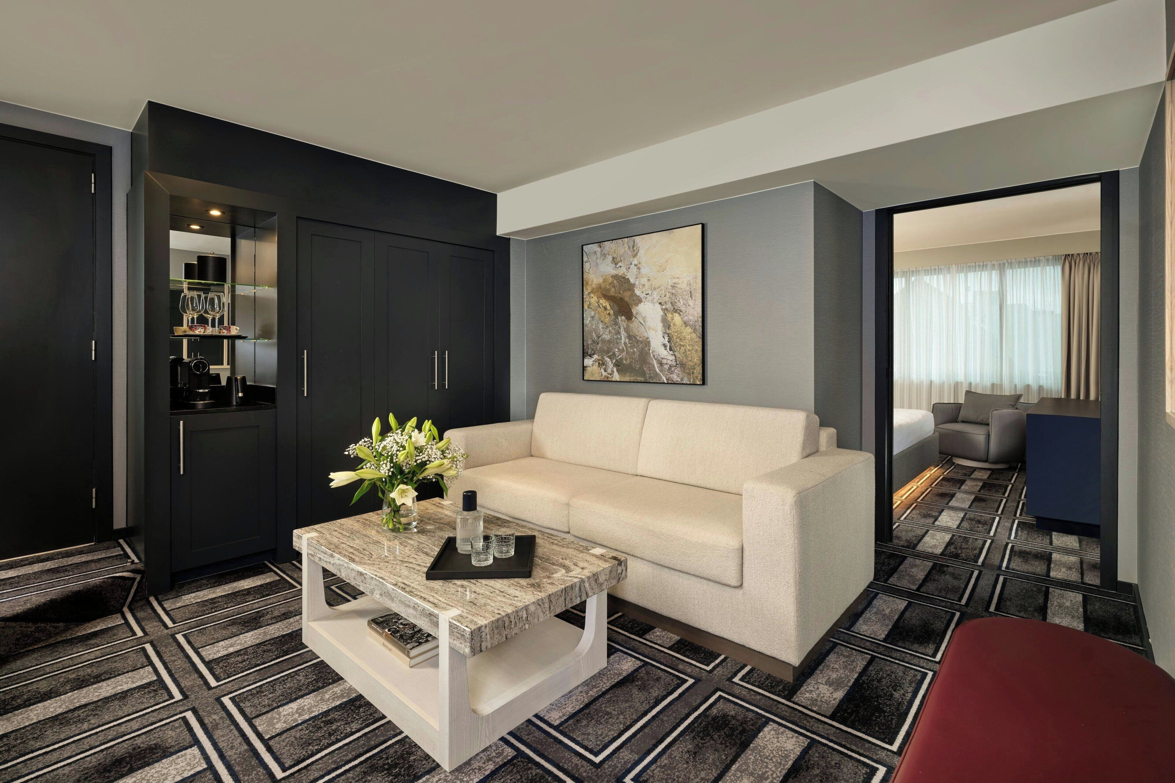 A modern hotel room with a sofa, a coffee table, a dining area with a chair, and a kitchen area with cabinets and a countertop.