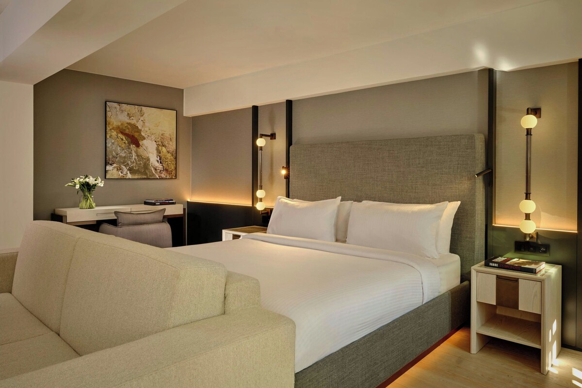 Una habitación de hotel moderna con una cama grande, un sofá, un escritorio y una mesita de noche con una lámpara.
