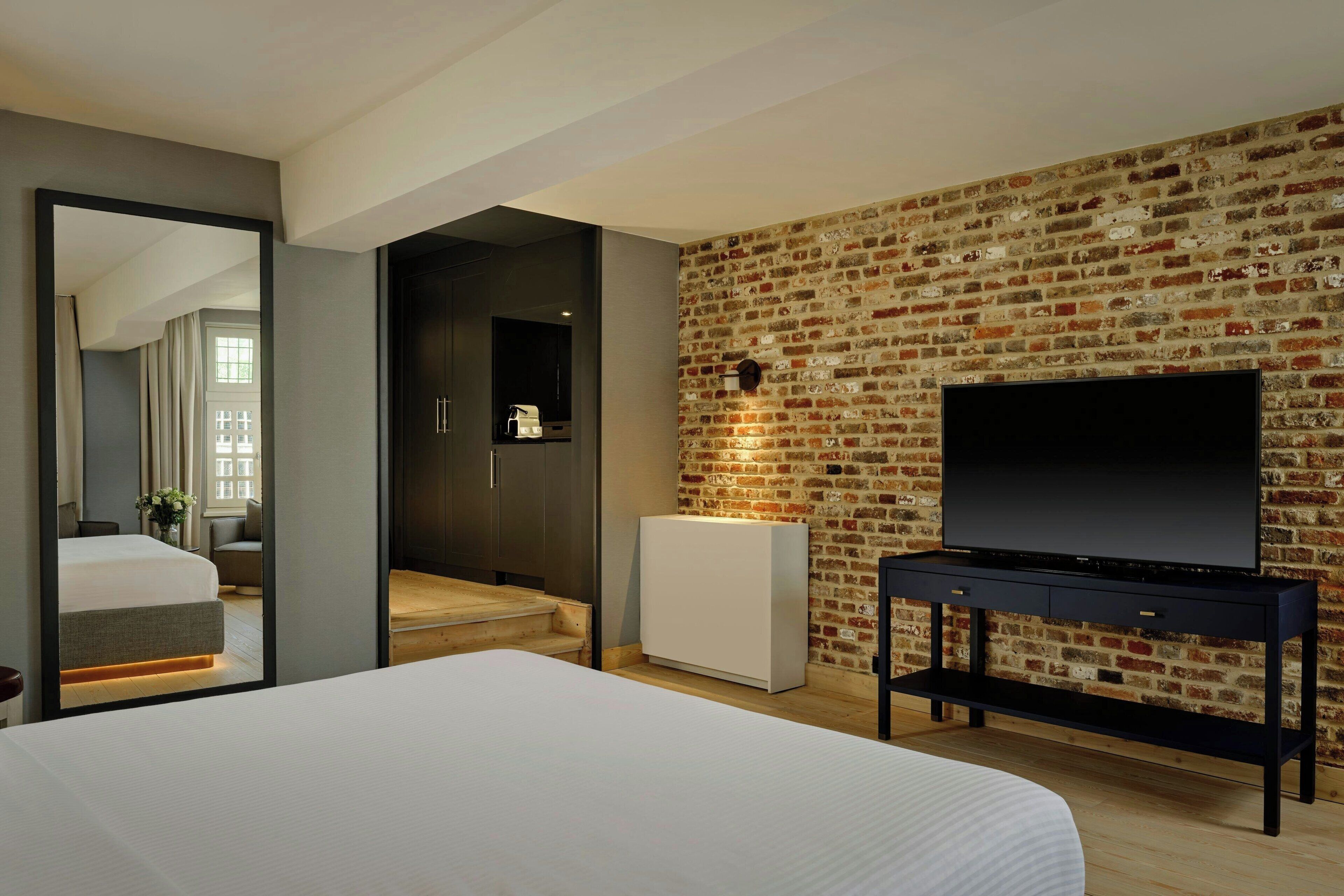 A modern hotel room with a large bed, a TV mounted on a brick wall, and a walk-in closet.