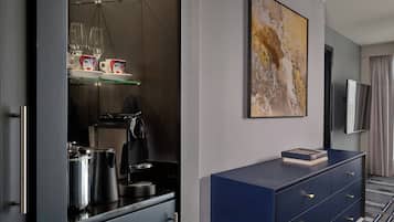 A modern hotel room with a kitchenette, a coffee machine, a glass shelf with cups, a large abstract painting, a flat-screen TV, and a blue dresser.
