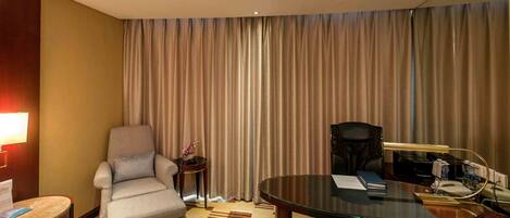 A hotel room with a large window, a desk with a lamp, a chair, and a small table.