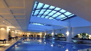 An indoor swimming pool with a glass ceiling, lounge chairs, and potted plants.