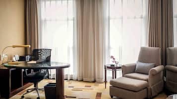 Una habitación de hotel moderna con una mesa de vidrio, una silla de oficina negra, un sofá color beige y una mesita auxiliar con un florero.