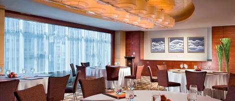 A hotel dining area with round tables, brown chairs, and a large window with sheer curtains.